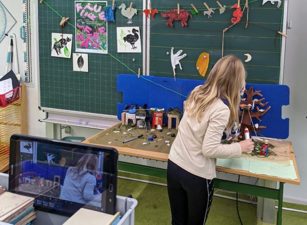 Zu sehen ist ein Klassenraum. Vor der Schultafel steht ein kin an einem Schultisch und baut mit gebastelten Gegenständen eine Szene für einen Stop Motion Film. Hinter ihr ist eine kamera zu sehen.
