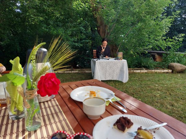 Zu sehen ist die Autorin Tina Pruschmann, die an einem Tisch mit weißer Tischdecke unter einem Baum sitzt. Sie ließt aus Ihrem Buch. Im Vordergrund ist ein Tisch mit Blumen, Tischdeck und Geschirr zu sehen. Auf einem Teller liegt eine Kuchengabel und Kuch