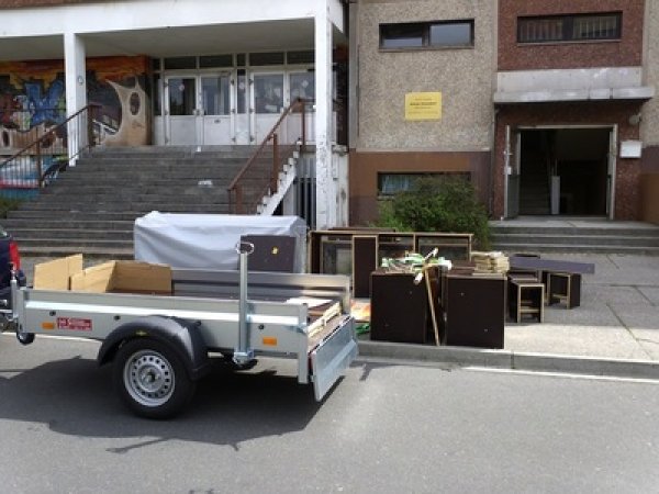 Zu sehen ist ein Autoanhänger vor einem Schulgebäude. Der Anhänger wird gerade ausgeladen. Auf dem Bürgersteig stehen verschieden dunkelbraune Holzkisten